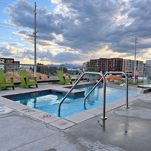 A rooftop pool with green chairs, a metal pool ladder, and a view of buildings and mountains in the background. - Downtown SLC Studio | King Bed + Free Parking (Salt Lake City)