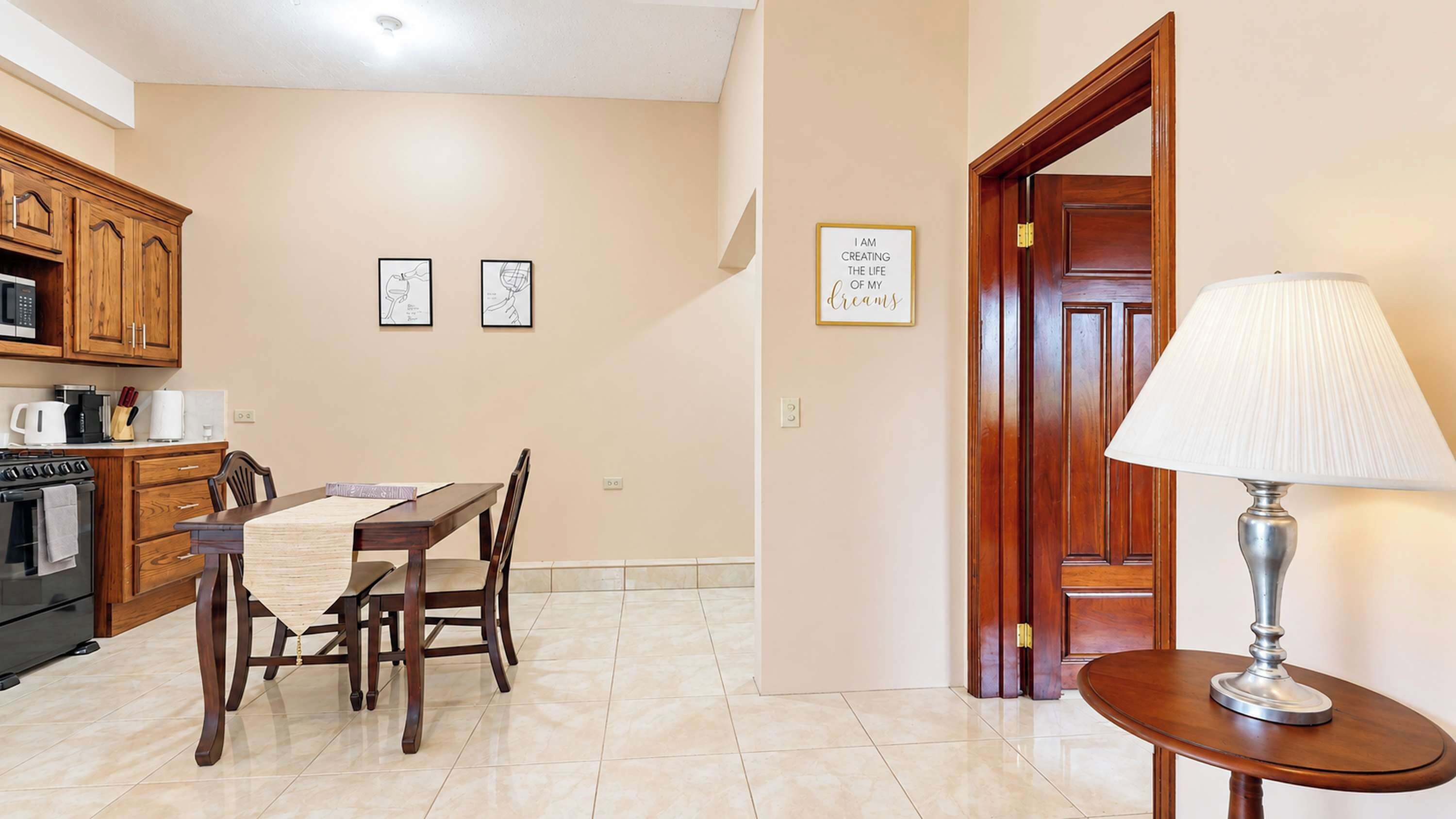 A kitchen with wooden cabinets, a dining table with chairs, a microwave, and a stove. There is a framed quote on the wall and a lamp on a side table.