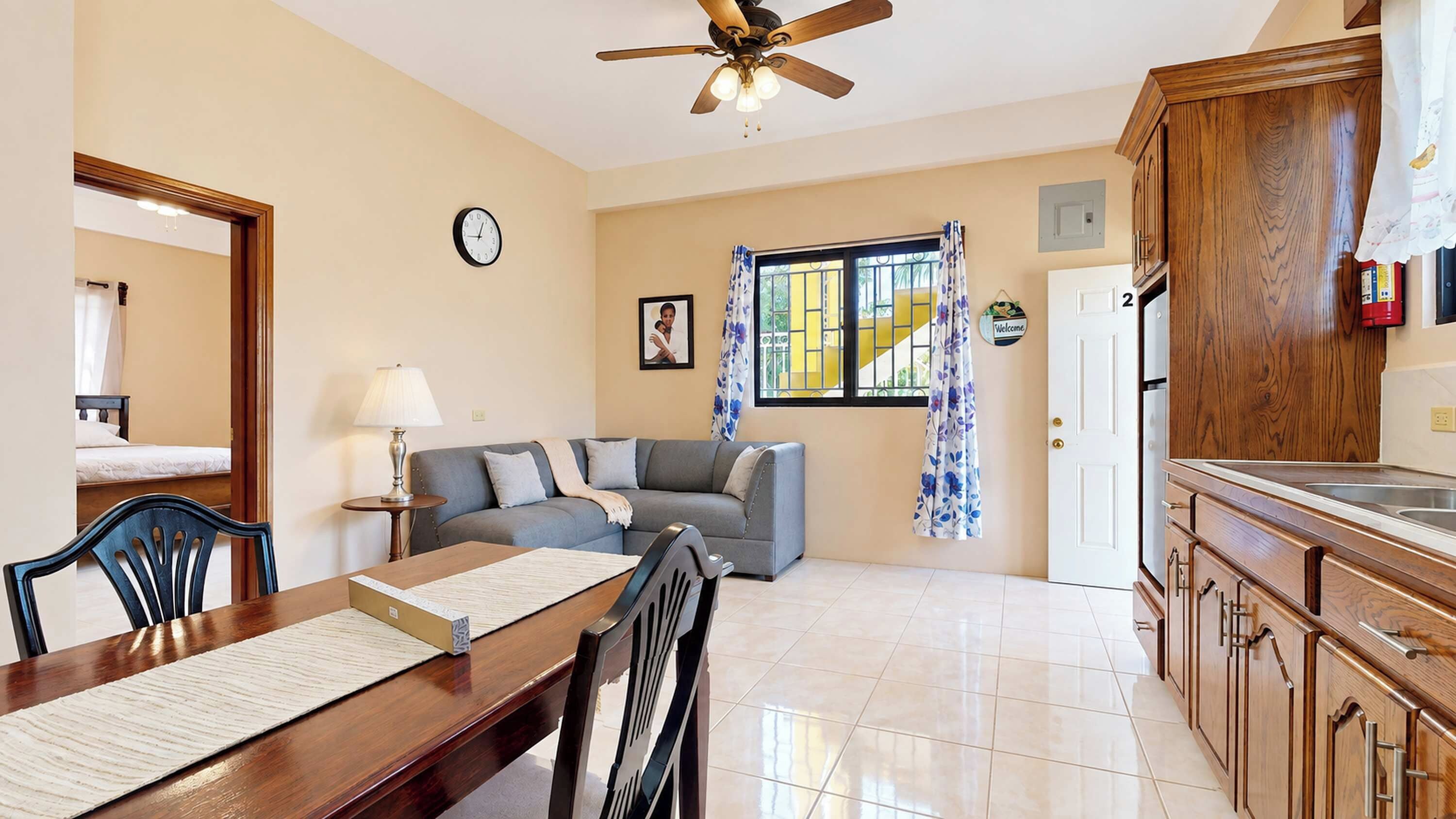 A living room with a sofa, a dining table with chairs, a kitchen area with wooden cabinets, a ceiling fan, and a window with curtains.
