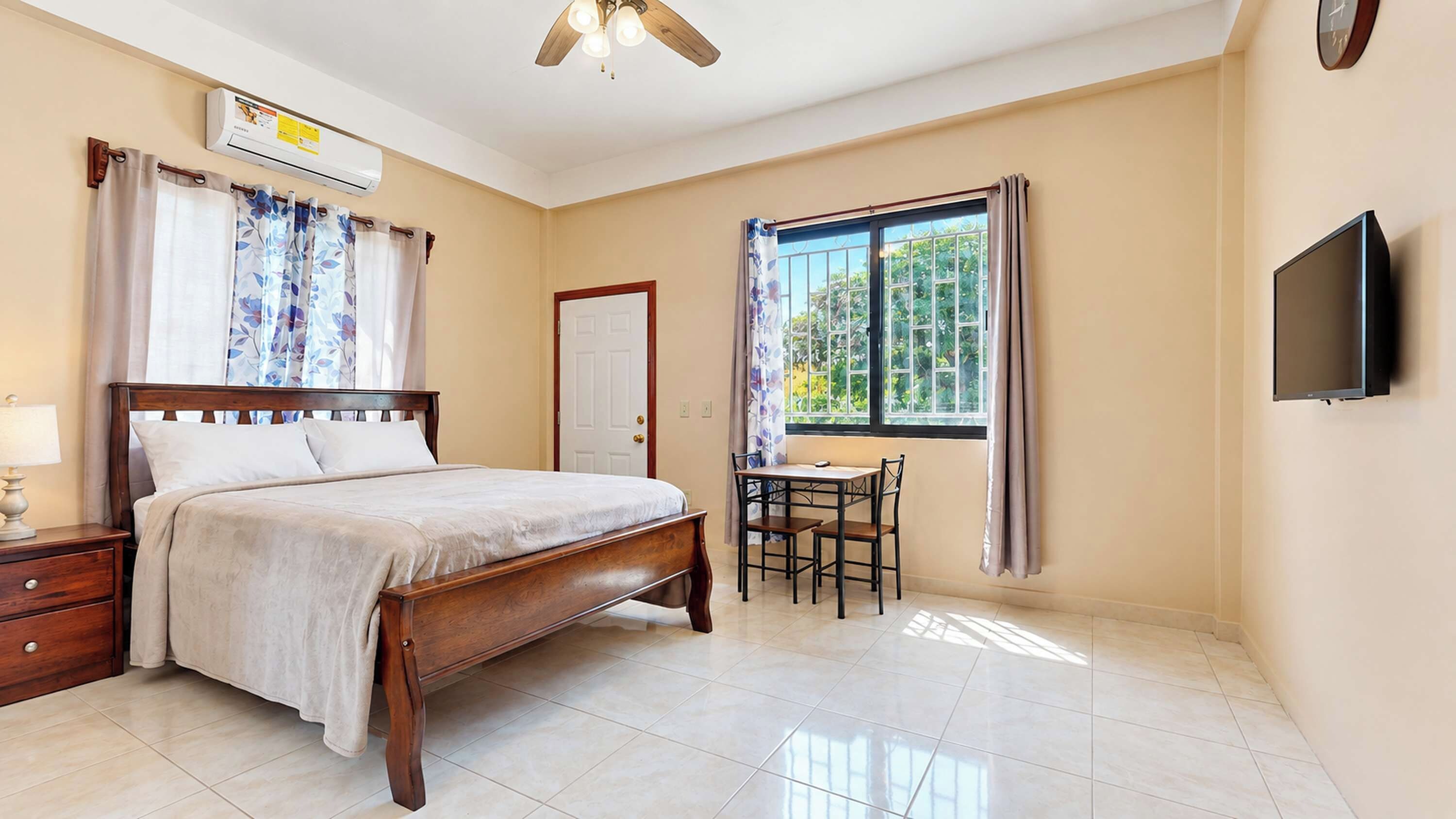 A bedroom with a bed, bedside table, lamp, air conditioning unit, ceiling fan, window with curtains, and a television mounted on the wall.