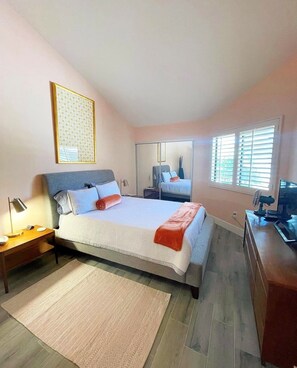 A bedroom with a bed, bedside tables, a desk, a chair, a TV, and a window with blinds. - Mid-Century Modern Desert Retreat With Golf Cart, Fairway Views & 48 Pools (Palm Desert)