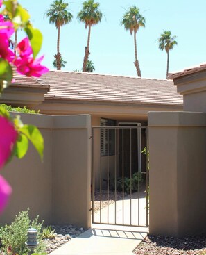 A gated entrance to a building with a tiled roof, surrounded by palm trees and a garden with rocks and plants. - Mid-Century Modern Desert Retreat With Golf Cart, Fairway Views & 48 Pools (Palm Desert)