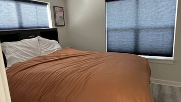 A bedroom with a bed, two windows with blinds, a headboard, and a wall-mounted artwork.