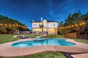 Casa de dos pisos con piscina, rodeada por una cerca de madera y luces colgantes.