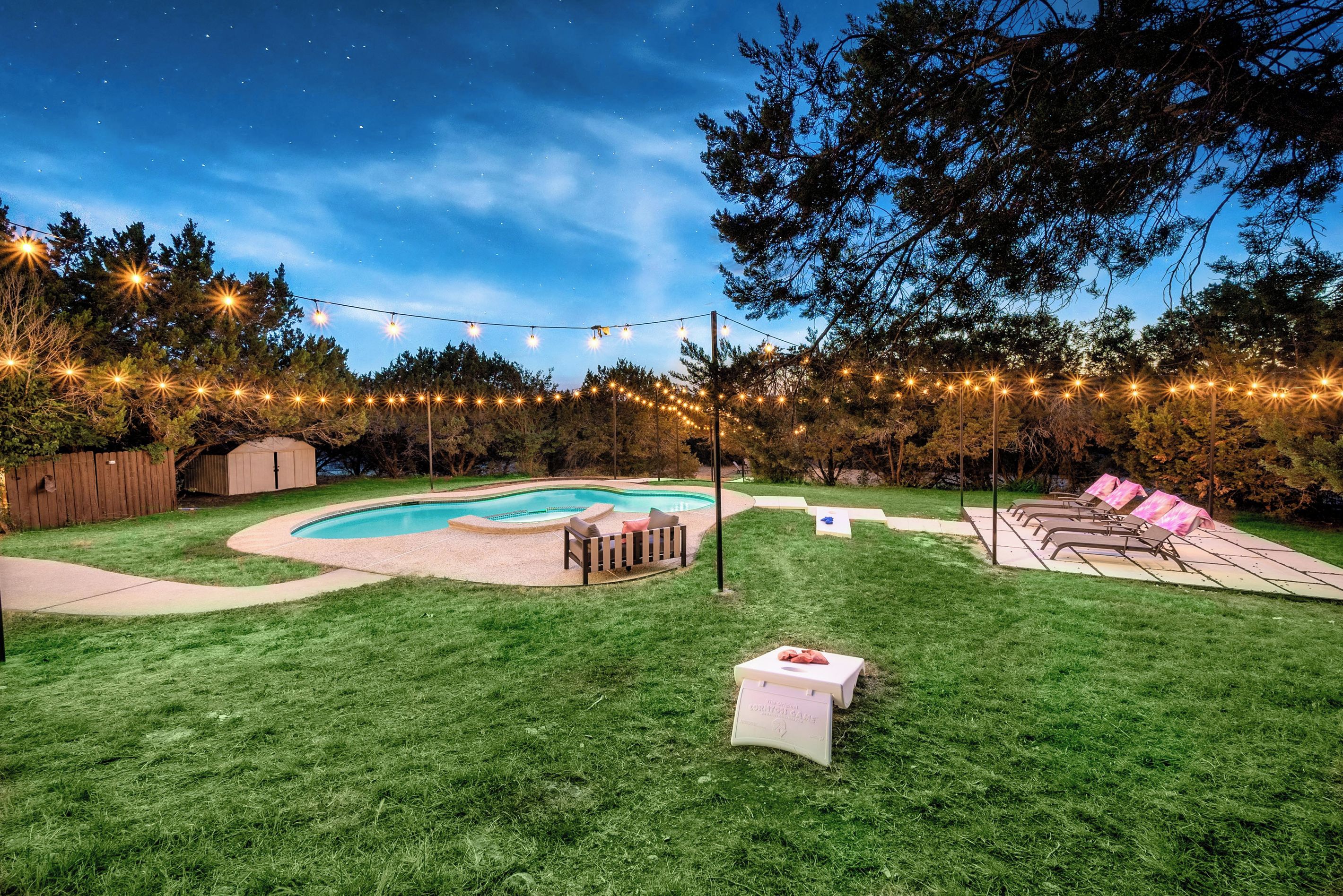 A backyard with a swimming pool, string lights, a wooden deck, and a grassy lawn.
