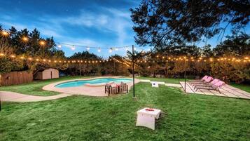 A backyard with a swimming pool, string lights, a wooden deck, and a grassy lawn.