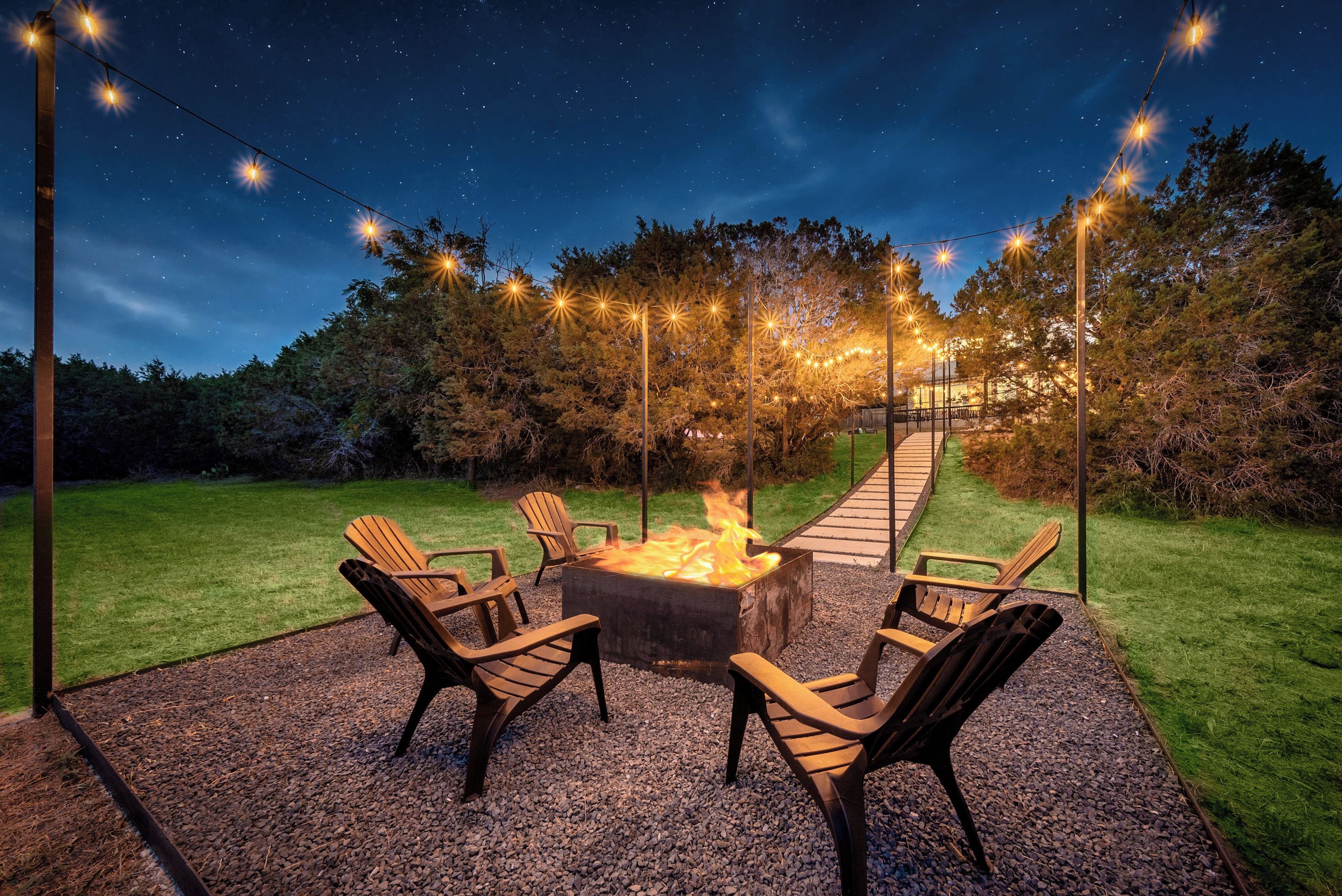 An outdoor seating area with a fire pit, surrounded by chairs and string lights against a night sky.