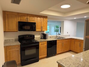 A kitchen with wooden cabinets, a black microwave, stove, and dishwasher, granite countertops, and a window providing natural light. - Beach, Golf, & Fitness Getaway, Newly Renovated. (Naples)