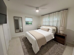 Un dormitorio con cama, mesitas de noche, un espejo, una ventana con cortinas y un ventilador de techo.