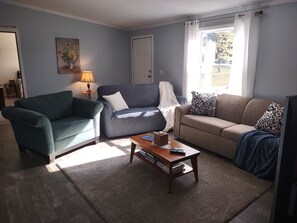 Ett vardagsrum med en blå fåtölj, en grå soffa, en beige soffa, ett träbord med böcker och en fjärrkontroll, och en tavla på väggen.