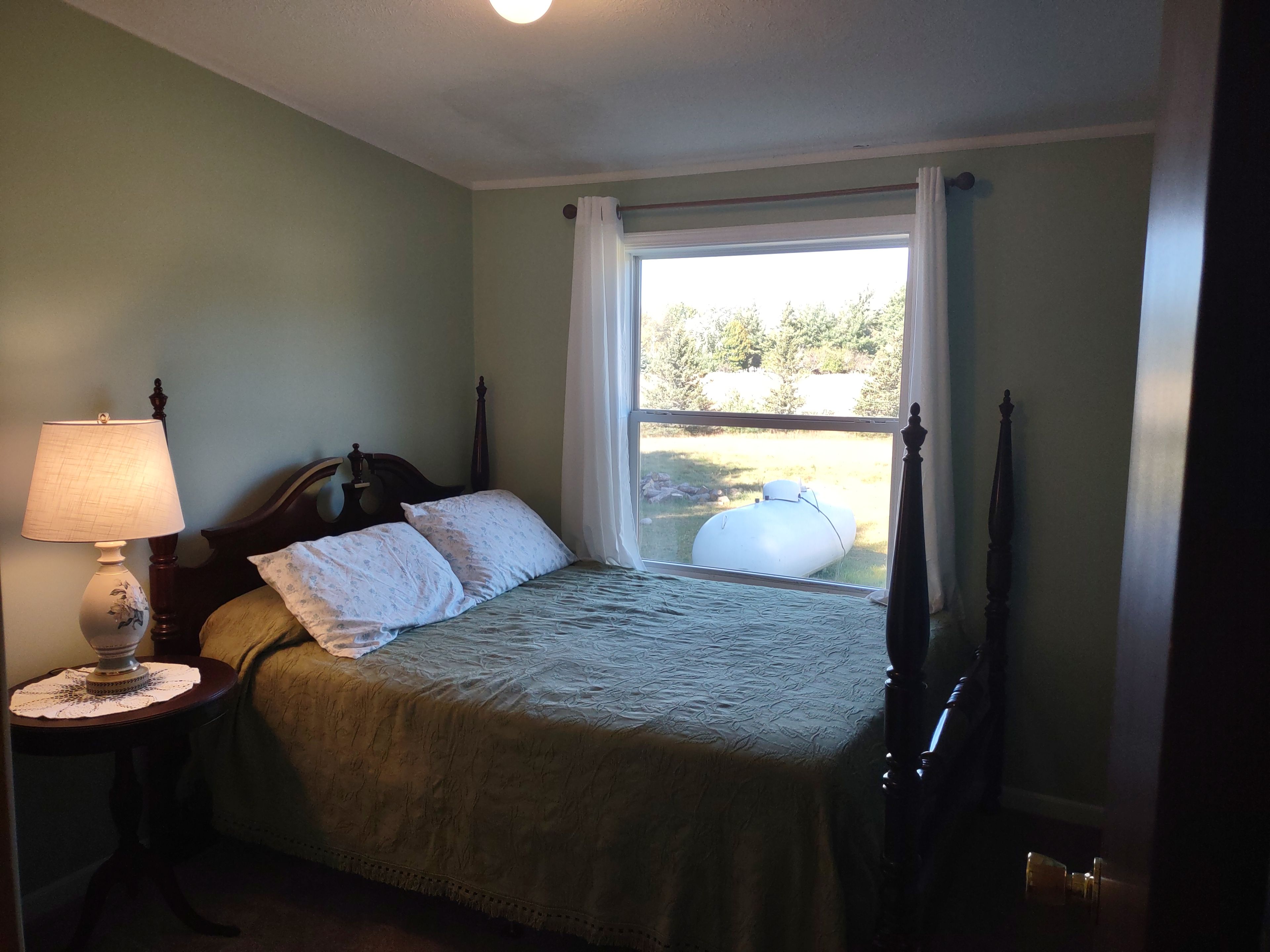 A bedroom with a bed, a nightstand with a lamp, a window with curtains, and a view of the outdoors.