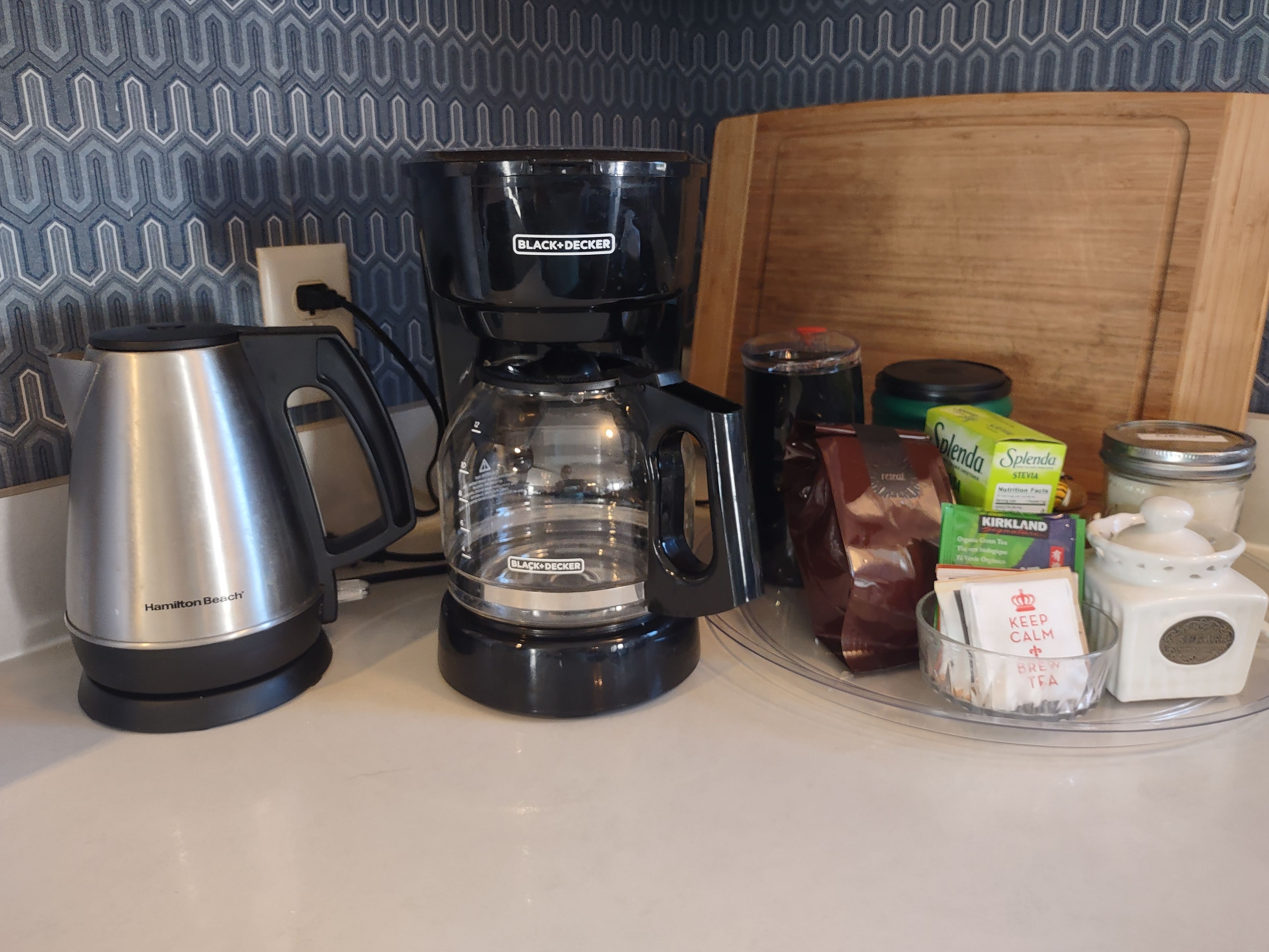 Un comptoir de cuisine avec une machine à café Hamilton Beach, une autre machine à café Black+Decker, un sachet de Splenda Stevia, une boîte de thé vert biologique Kirkland et un récipient Keep Calm & Been TFA.