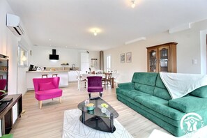 A modern living room with a green sofa, a pink armchair, and a purple armchair. There is a glass coffee table with a vase and a remote control. The room has wooden flooring and white walls. - Léonin's House - BAUD (BAUD)