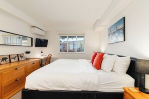 A modern bedroom with a bed, a desk, a chair, a dresser, a television, and a window with a view of a house and trees. - Steps to Gondola | Upper Village Loft | BBQ Deck | Fitness Room (Whistler)