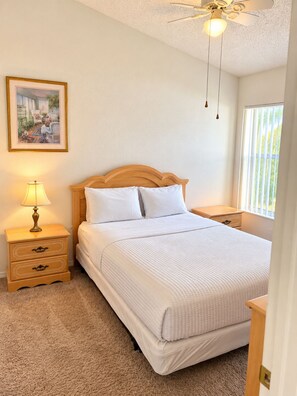 A bedroom with a bed, a nightstand with a lamp, a ceiling fan, a window with blinds, and a framed picture on the wall. - Perfect pool house with waterfront for the best Disney experience in Kissimmee. (Kissimmee)