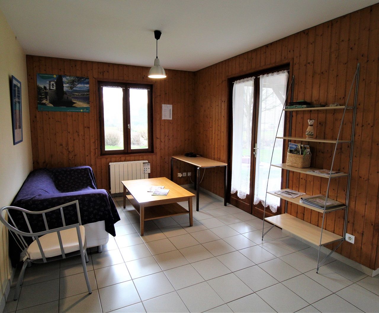 A room with a bed, a table, a chair, a radiator, a shelf, and a window.