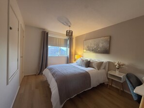 A bedroom with a bed, a nightstand, a lamp, a chair, a painting on the wall, and a window with curtains. - Cosy & Modern 2 bed house |Close to City Centre| Driveway (Nottingham)