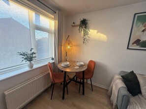 Un comedor con una mesa dispuesta para dos, una ventana con cortinas, una planta colgante y un cuadro en la pared.