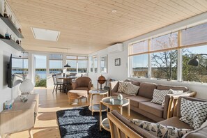 A spacious living room with a large window overlooking a body of water, a cozy seating area, and a dining space. - Cozy holiday home with private jetty (Randers NØ)
