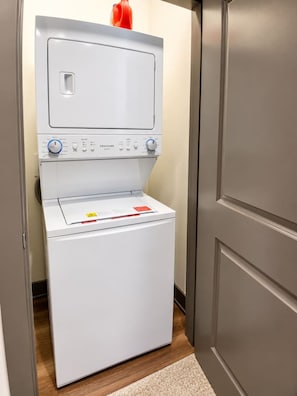 A compact, white front-loading washing machine in a hallway with a wooden floor and a beige carpet. - Modern Stadium Retreat | Gym + Rooftop Lounge (Atlanta)