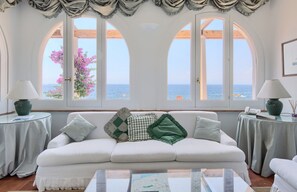 A white sofa with multiple cushions, a glass coffee table, and a view of the sea through large arched windows. - Apartment with Sea View a few steps from the beach (Massa Lubrense)