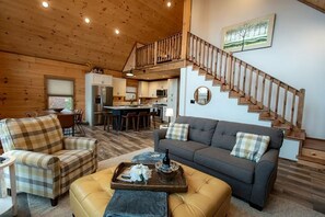 A spacious living area with a sofa, armchair, and ottoman, a wooden staircase, and a kitchen in the background. - Cookie Top Mountain Chalet (Caldwell)