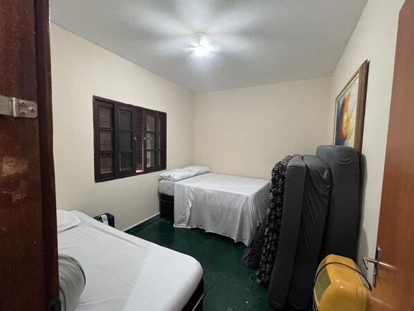 A small, compact room with two single beds, a window with shutters, a ceiling fan, and a door. - Country house with heated pool and gourmet area in an apartment in Goiânia. (Aparecida de Goiânia)