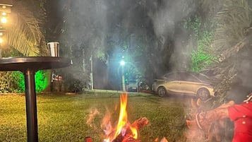 Un barbecue avec des flammes, une voiture et une personne vêtue de rouge dans un jardin, de nuit.