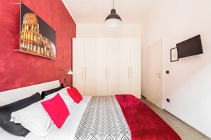 A bedroom with a bed, a red wall, a patterned rug, a white wardrobe, and a flat-screen TV. - Riari 59, Rome - Trastevere (Roma)