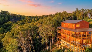 A two-story wooden cabin with a balcony, surrounded by dense forest. - Heaven's Grace Pool Cabin by Avantstay Smokies Retreat Pool + Hot Tub + Game Room + Theater (Sevierville)