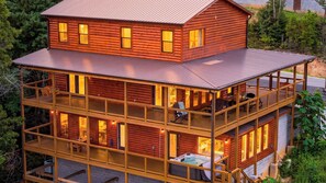 A multi-level log cabin with a hot tub on the ground floor, outdoor seating on the upper level, and a view of the surrounding forest. - Heaven's Grace Pool Cabin by Avantstay Smokies Retreat Pool + Hot Tub + Game Room + Theater (Sevierville)