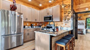 A modern kitchen with stainless steel appliances, wooden cabinets, a marble countertop, and a central island with bar stools. - Heaven's Grace Pool Cabin by Avantstay Smokies Retreat Pool + Hot Tub + Game Room + Theater (Sevierville)