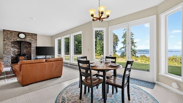 A modern living room with a fireplace, a dining area with a table set for four, and large windows offering a view of the outdoors.