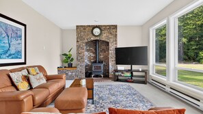 A living room with a brick fireplace, a leather sofa, a flat-screen TV, and a large window offering a view of the outdoors. - The Eagle's Nest by Avantstay 6-acre Oak Harbor Retreat Fire Pit Mt. Baker + Ocean Views (Oak Harbor)