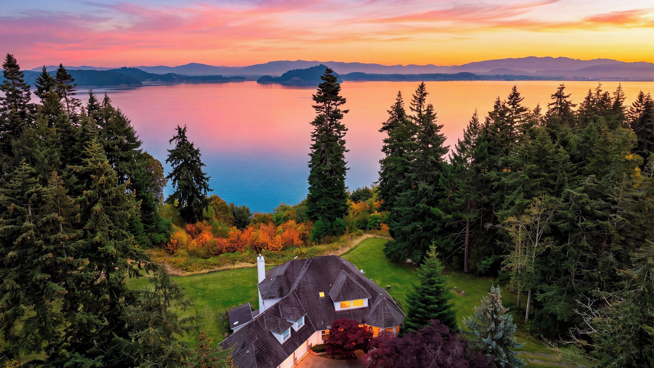 A house with a sloped roof, surrounded by dense forest, overlooking a calm lake with mountains in the distance.