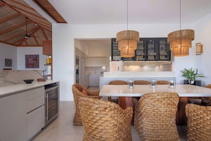 A modern kitchen with a central island, built-in appliances, and woven chairs. - Villa La Perla: 4BR Four Seasons Villa w/ Private Pool, Chef & Resort Access (Punta Mita)