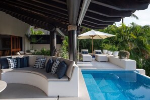 Zona junto a la piscina con un sofá blanco curvo, una piscina y un área de descanso cubierta con sombrillas.