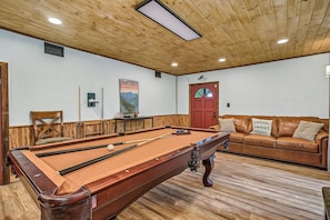 A pool table in a room with wooden flooring, a red door, and a brown leather couch with a cushion. - Halfway to Heaven · Hot Tub, Game Room, Mtn View (Gatlinburg)