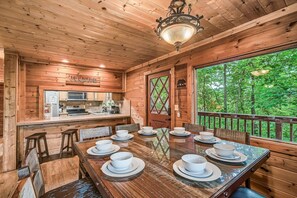 A wooden cabin interior with a dining table set for six, a kitchen area, and a large window overlooking greenery. - Halfway to Heaven · Hot Tub, Game Room, Mtn View (Gatlinburg)