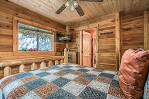 A cozy cabin room with a quilted bedspread, a wooden bench, a television, and a window with a view of greenery. - Halfway to Heaven · Hot Tub, Game Room, Mtn View (Gatlinburg)