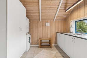 A compact kitchen with wooden walls, white cabinets, a built-in washing machine, and a window with a view of trees. - Cosy holiday home near Ebeltoft and the beach (Ebeltoft)