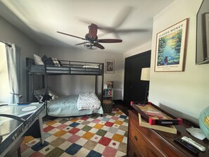 Un dormitorio con literas, un escritorio, un ventilador de techo, una alfombra colorida, una estantería y un cuadro enmarcado.