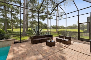 A sunlit patio with a pool, a seating area with a sofa and chairs, and a view of a golf course through large windows. - Elegant Country Club House with Pool View of the 14th Hole (Orange Park)