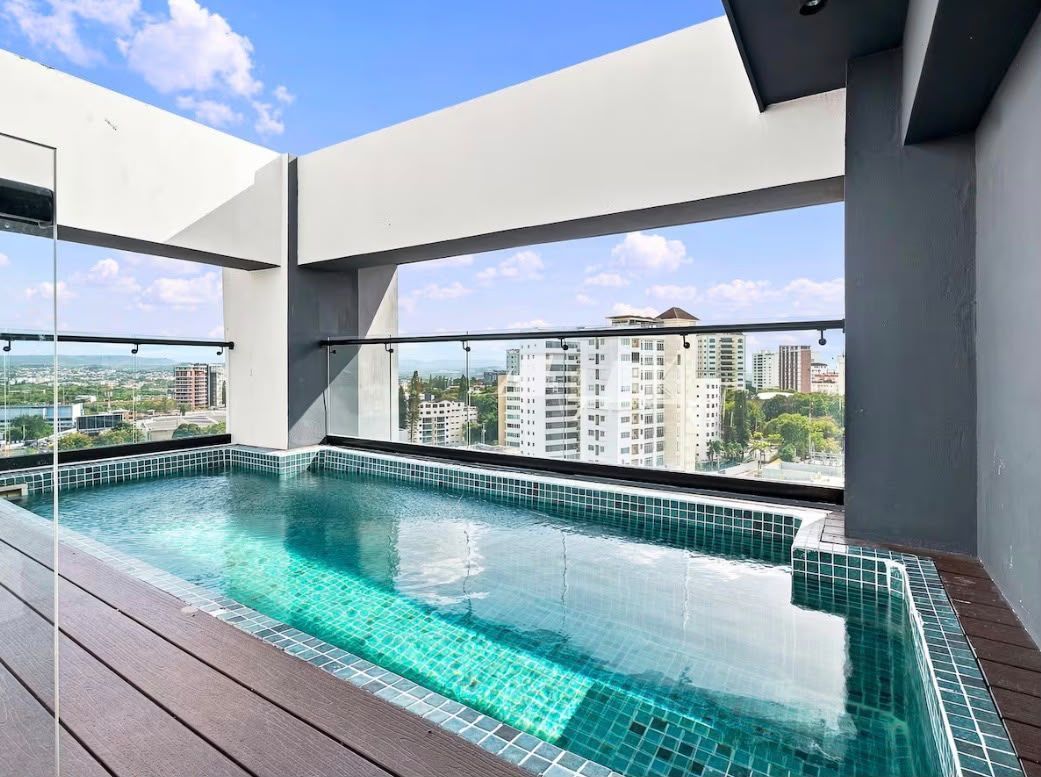A rooftop swimming pool with clear blue water, surrounded by a wooden deck and white walls.
