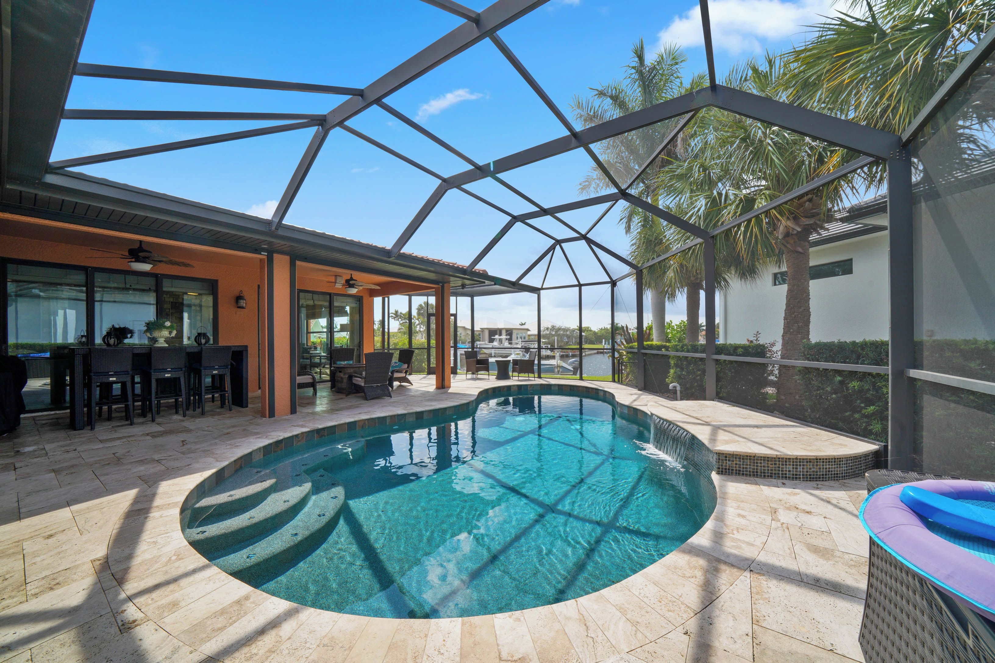 A swimming pool with a glass enclosure, outdoor seating, and a view of palm trees.