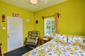 Een kamer met een bed, een stoel, een raam met gordijnen, een vluchtroutebord en een brandblusser.