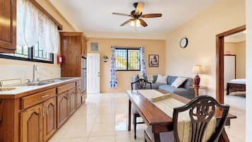 A well-lit kitchen with wooden cabinets, a dining table with chairs, a ceiling fan, and a view to a living area with a sofa and a lamp.