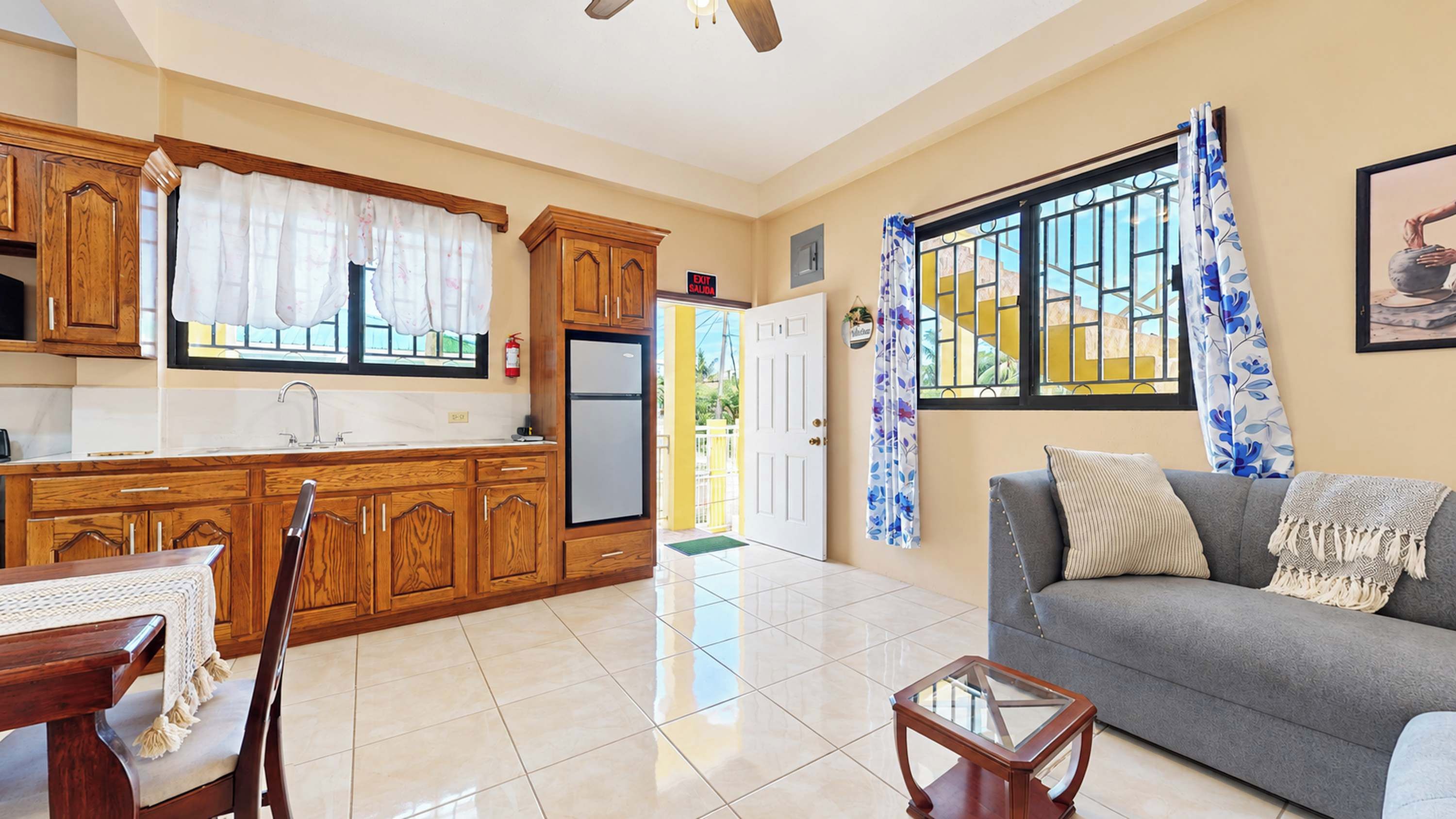 A living room with a grey sofa, a wooden coffee table, and a kitchen with wooden cabinets and a refrigerator.