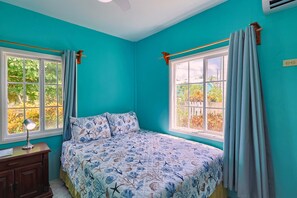 Une chambre avec un mur de couleur turquoise, un lit recouvert d’une courtepointe à motifs floraux, deux fenêtres avec des rideaux, une table de chevet avec une lampe et une commode en bois.