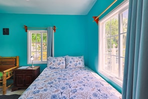 A room with teal walls, a bed with a floral bedspread, a wooden desk, and a window with curtains. - Central Caye Caulker Apartment w/ AC, Wi-Fi & Kitchen | Unit 2 (Caye Caulker)